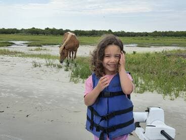 A joyful child in a life jacket smiles while a wild horse grazes nearby, showcasing a delightful moment of human-animal interaction in a natural setting.