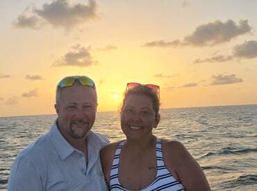 A couple smiles together against a stunning sunset backdrop on the water, capturing a perfect moment of romance and celebration.
