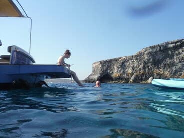 A woman sits on the edge of a boat while another person enjoys a swim in clear waters near a rocky coastline, capturing the essence of a fun day on the water.