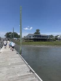 A group of people strolls along a wooden dock, enjoying a sunny day by the waterfront, with scenic views of the surrounding area.