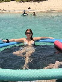 A woman relaxes on a floating mat in crystal-clear waters while friends enjoy the beach in the background — a perfect day for fun and relaxation in the sun.