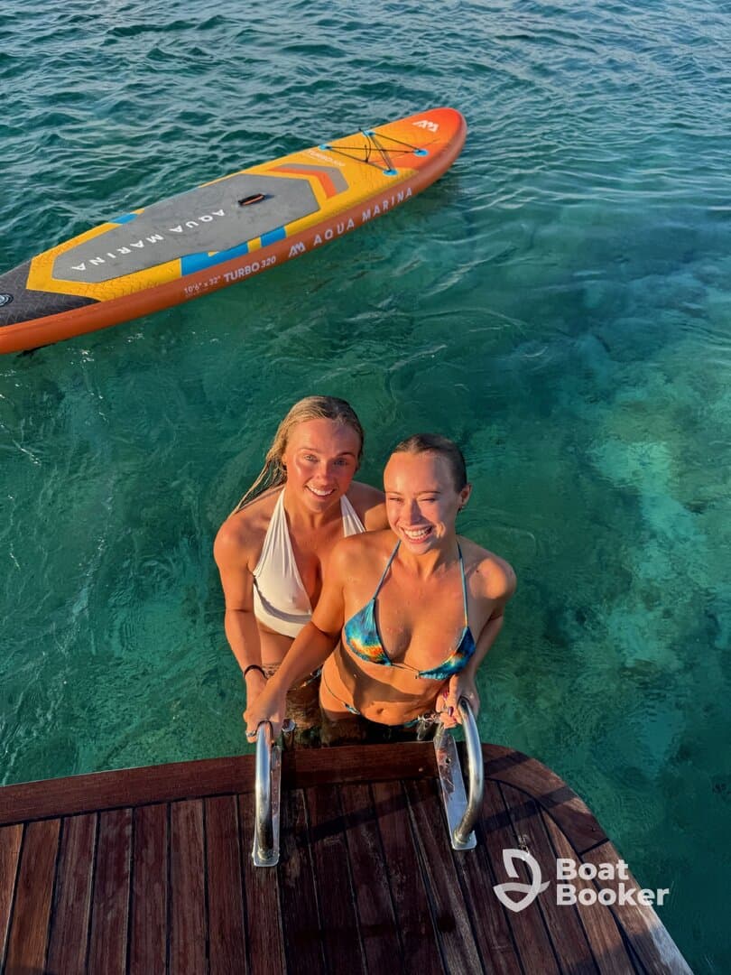 Two friends enjoy a refreshing swim and pose for a photo while holding onto the ladder of a boat, with a paddleboard nearby — a perfect moment of fun in the sun.