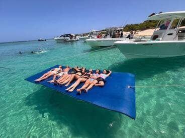 A lively group enjoys a sunny day on a floating mat in crystal-clear waters, surrounded by boats — perfect for summer fun and beach parties.
