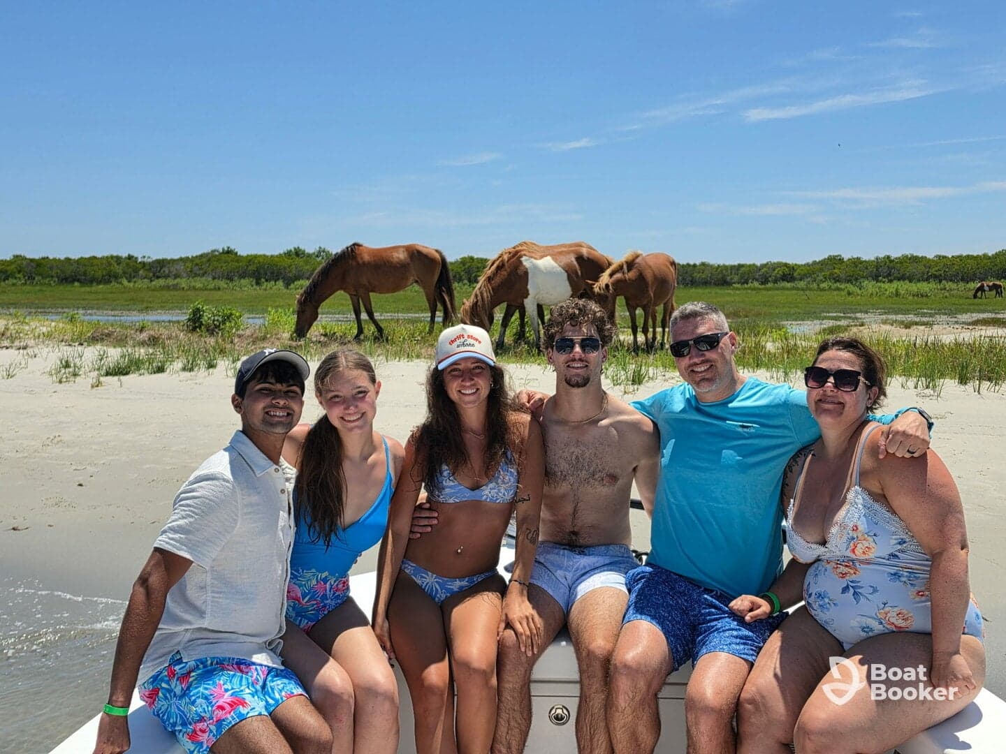 A joyful group poses on a boat with wild horses grazing in the background, capturing a memorable beach day filled with fun and adventure.