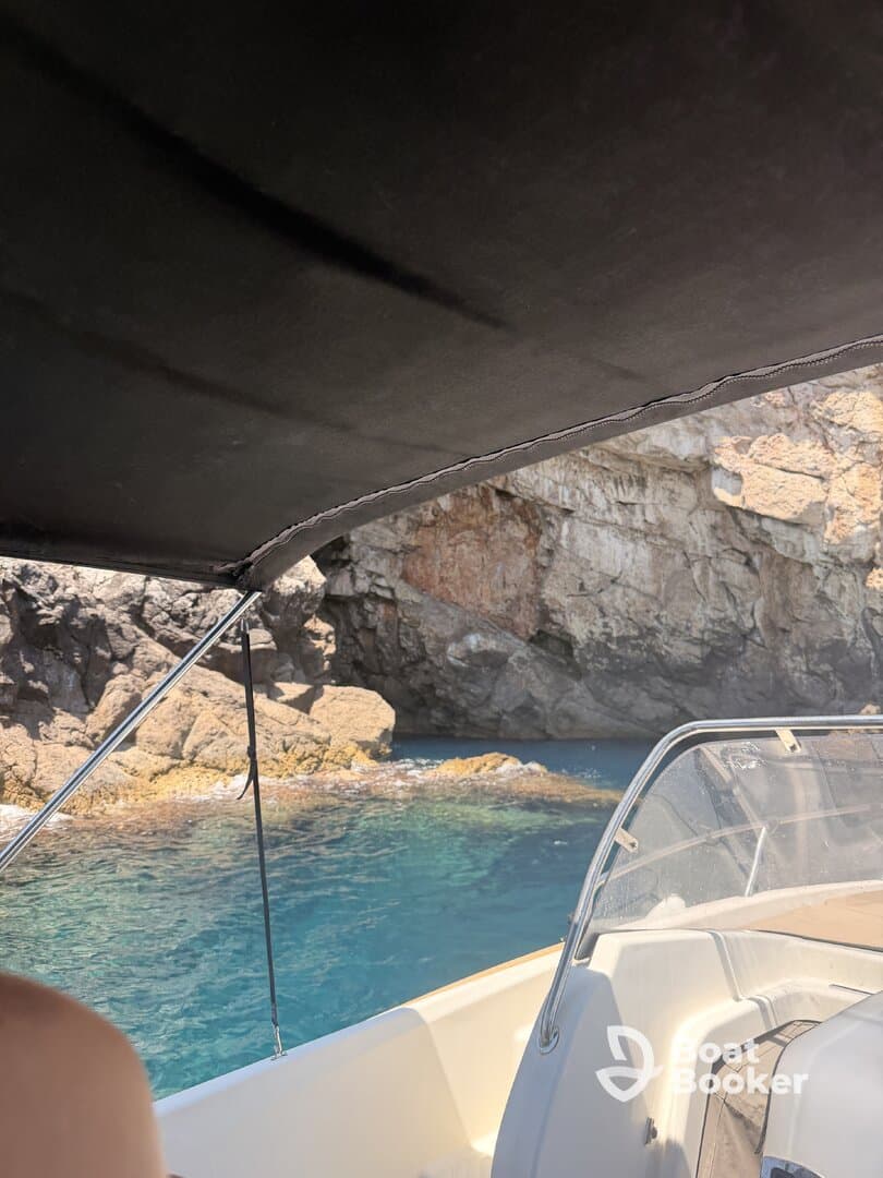 A view from inside a boat's cabin, showcasing the helm and shaded area while navigating near rocky cliffs and clear blue waters.
