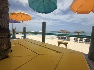 A view from a beachside table with colorful umbrellas and a glimpse of the ocean, creating a relaxed atmosphere. The focus is unclear, making it difficult to identify specific details.