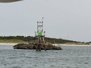 An unclear view of a lighthouse structure on a rocky outcrop, surrounded by water and greenery in the background.