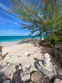 A serene beach scene featuring a person standing near stacked rocks, with turquoise waters and a clear blue sky in the background — perfect for relaxation and nature appreciation.