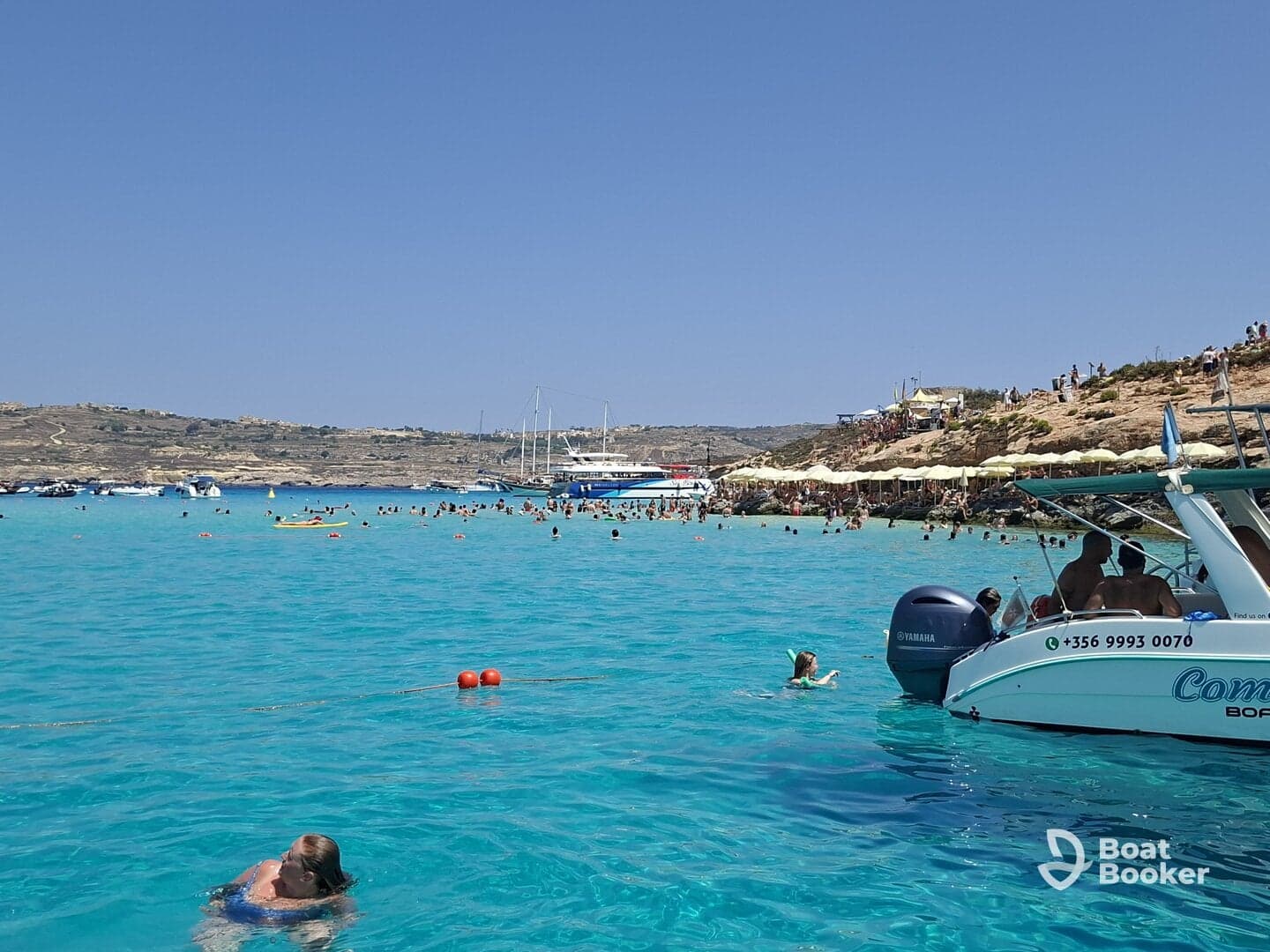 A vibrant scene at a popular beach with people swimming in crystal-clear turquoise waters, enjoying a sunny day with boats anchored nearby.