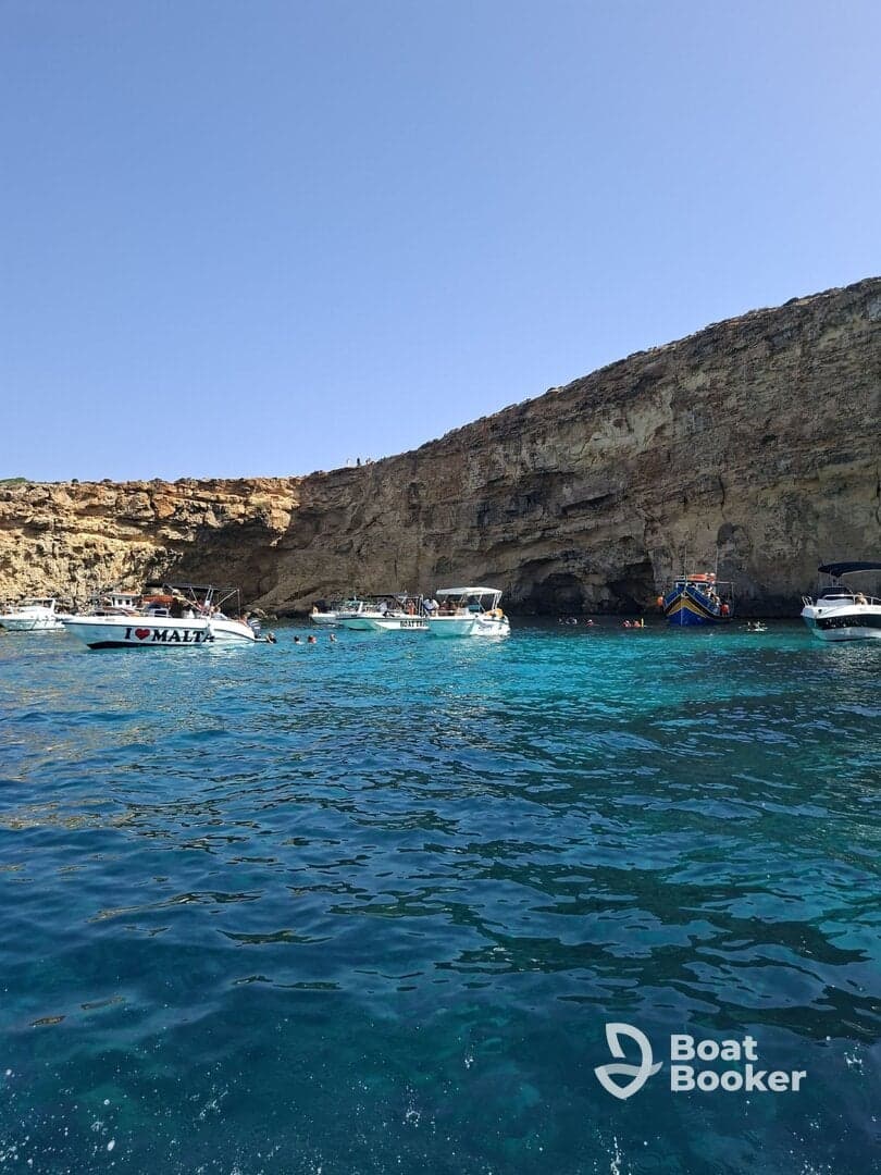A vibrant scene in Malta showcasing multiple boats anchored near a stunning rocky coastline, with people enjoying the clear turquoise waters — a perfect destination for swimming and exploration.