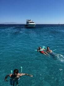 A lively group enjoys a sunny day swimming in crystal-clear waters, with a yacht anchored nearby — perfect for a fun day on the water.