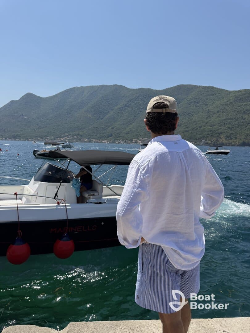A person stands by the water, gazing at a boat named 'Martinello' amidst a stunning mountainous backdrop — a perfect scene for a summer day on the water.
