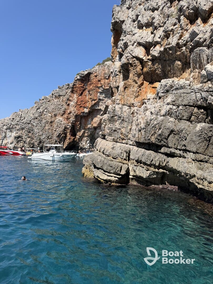 A picturesque view of a rocky coastline with boats anchored nearby, while a swimmer enjoys the clear blue waters — a perfect spot for a summer adventure.