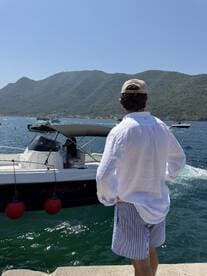 A person stands by the water, gazing at a boat named 'Martinello' amidst a stunning mountainous backdrop — a perfect scene for a summer day on the water.