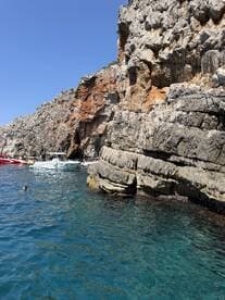 A picturesque view of a rocky coastline with boats anchored nearby, while a swimmer enjoys the clear blue waters — a perfect spot for a summer adventure.