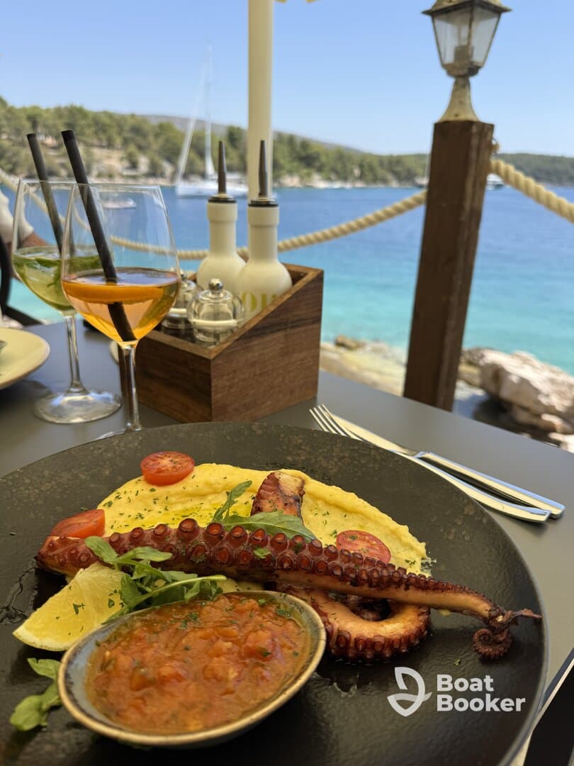 A beautifully plated dish featuring grilled octopus, accompanied by fresh tomatoes and a savory sauce, set against a stunning coastal backdrop — perfect for enjoying gourmet meals while on a boat trip.