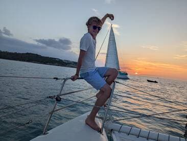 A young adult enjoys the sunset while sailing, striking a playful pose on the bow of the yacht — a perfect moment for adventure and relaxation on the water.