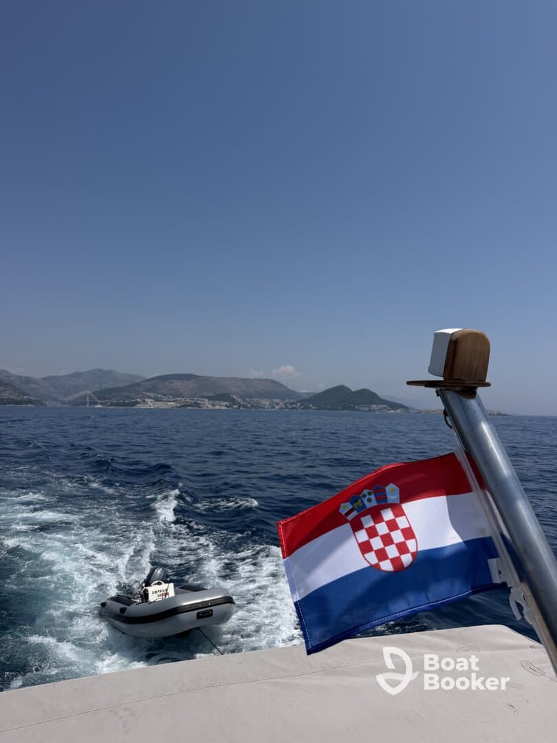 A scenic view of a boat in motion, showcasing the Croatian flag and a tender in tow against a backdrop of beautiful coastal mountains.