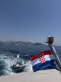 A scenic view of a boat in motion, showcasing the Croatian flag and a tender in tow against a backdrop of beautiful coastal mountains.