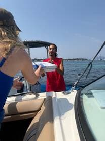A woman hands over a pizza box to a man on a boat, capturing a fun food delivery experience on the water — perfect for enjoying meals while cruising.