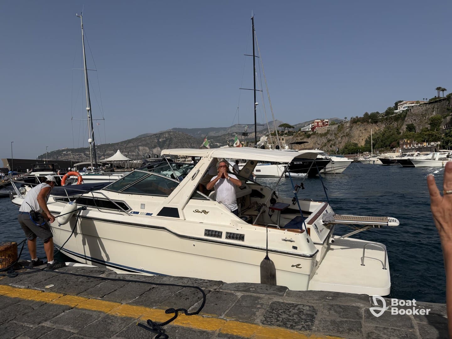 Two individuals are seen interacting on a boat while docking at a bustling harbor, showcasing the vibrant atmosphere of maritime life.