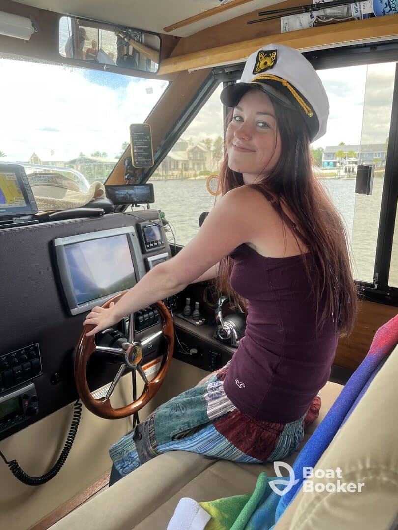 A young woman sits at the helm of a boat, wearing a captain's hat and smiling, showcasing the cozy interior of the vessel.