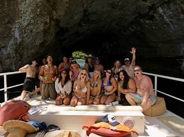 A lively group of friends celebrates aboard a boat near a stunning cave, enjoying drinks and creating unforgettable memories in a picturesque setting.