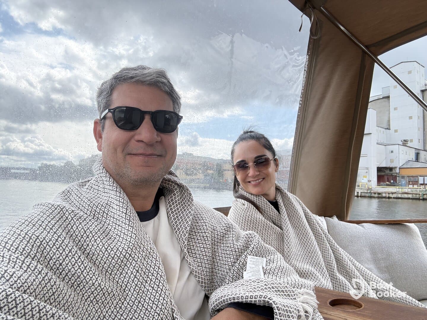 A couple enjoys a cozy moment on a boat, wrapped in blankets while taking in the scenic waterfront views — perfect for a relaxing day on the water.