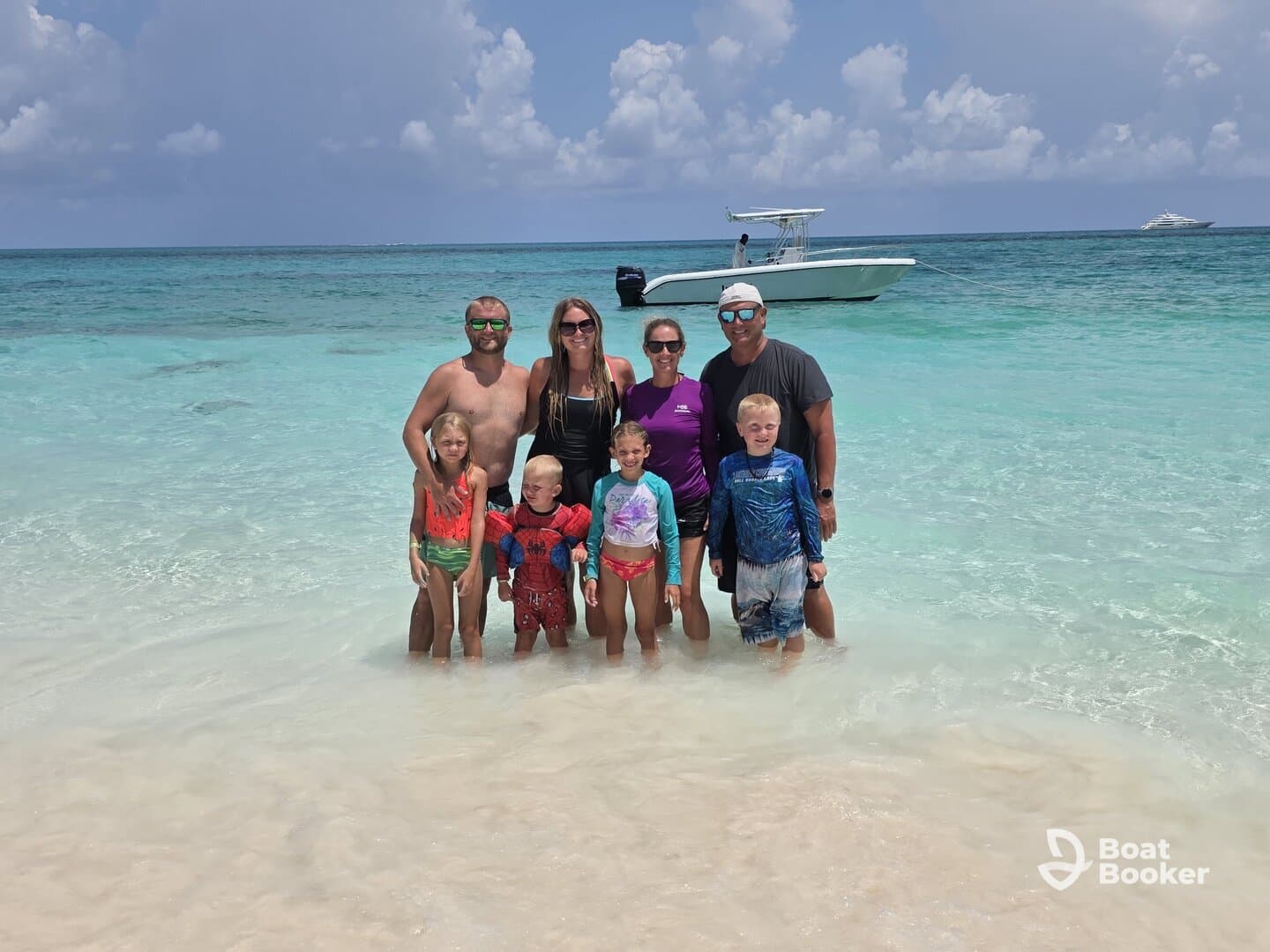 A joyful family poses together in shallow waters at the beach, with a boat anchored nearby — a perfect moment capturing fun and relaxation on a sunny day.