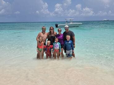 A joyful family poses together in shallow waters at the beach, with a boat anchored nearby — a perfect moment capturing fun and relaxation on a sunny day.