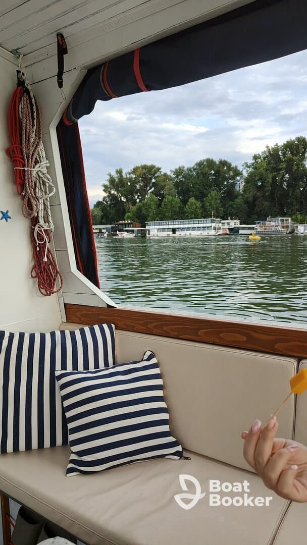 A cozy interior shot of a boat featuring striped cushions and a hand holding a snack, with a scenic view of the water and other boats in the background.