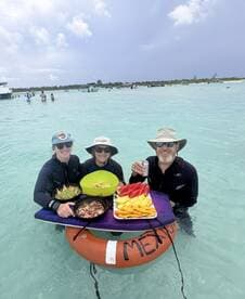 A cheerful group enjoys a delicious spread of fresh fruits and snacks while wading in the clear waters, perfect for a fun day of snorkeling and relaxation.