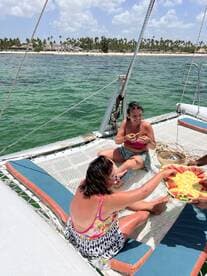 Two friends enjoy fresh fruit on the deck of a catamaran, soaking up the sun and the beautiful beach backdrop — a perfect moment for relaxation and fun on the water.