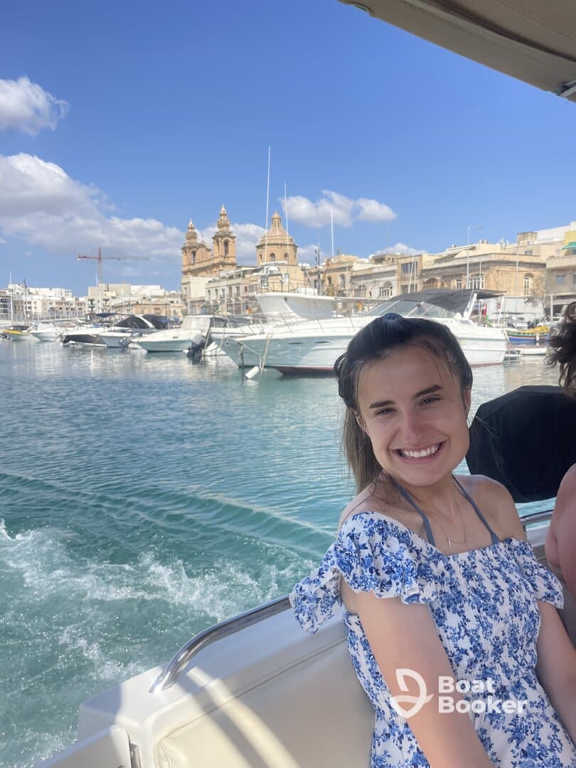 A young woman smiles while enjoying a boat ride in Malta, with a picturesque cityscape and clear waters in the background — a perfect snapshot of a memorable day on the water.
