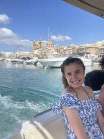 A young woman smiles while enjoying a boat ride in Malta, with a picturesque cityscape and clear waters in the background — a perfect snapshot of a memorable day on the water.