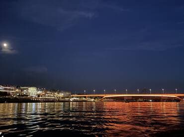 A serene night view of a cityscape illuminated by moonlight, with reflections shimmering on the water — a perfect backdrop for evening boat cruises.