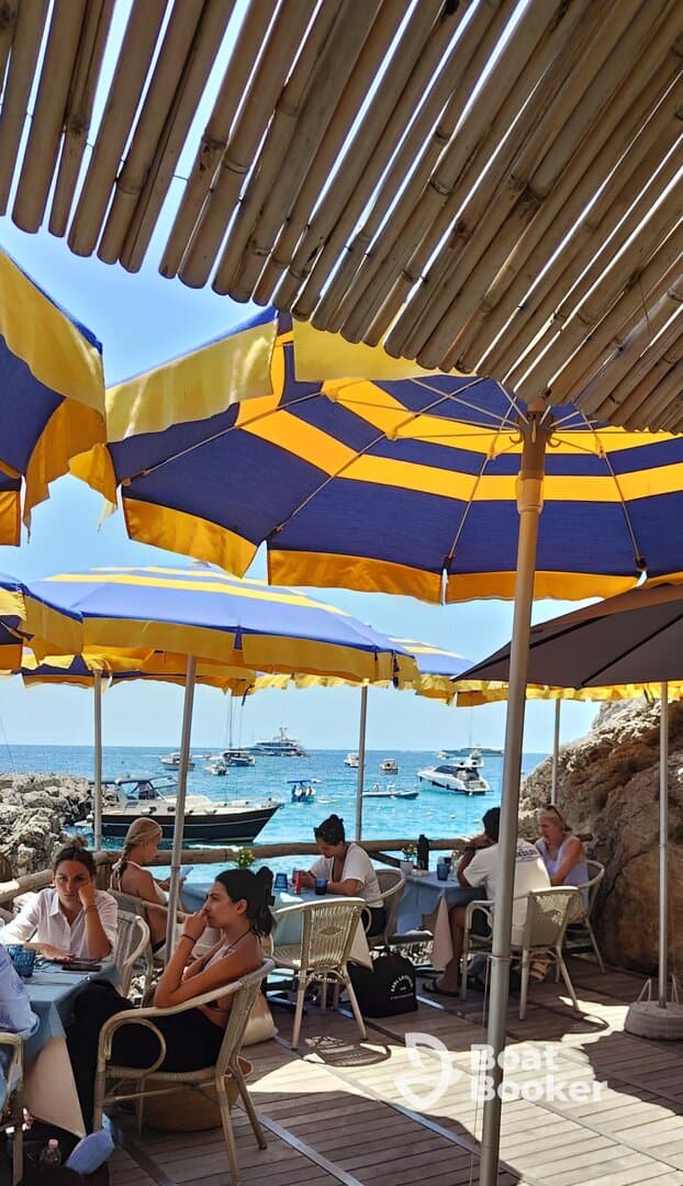 Guests enjoy a relaxing meal under colorful umbrellas at a seaside dining area, with boats visible in the background — a perfect spot for enjoying the coastal atmosphere.