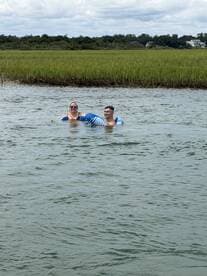 A couple enjoys a relaxing swim in the water, floating on inflatable loungers amidst a scenic backdrop of marshland — perfect for summer fun and water adventures.