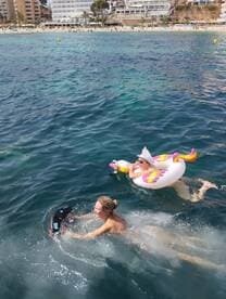 Two friends enjoy a sunny day in the water, one on a colorful inflatable while the other swims, capturing the essence of fun beach activities.