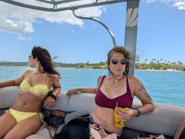 Two friends enjoy a sunny day on a boat, relaxing in their bikinis while taking in the beautiful waterfront scenery — perfect for a fun day out on the water.