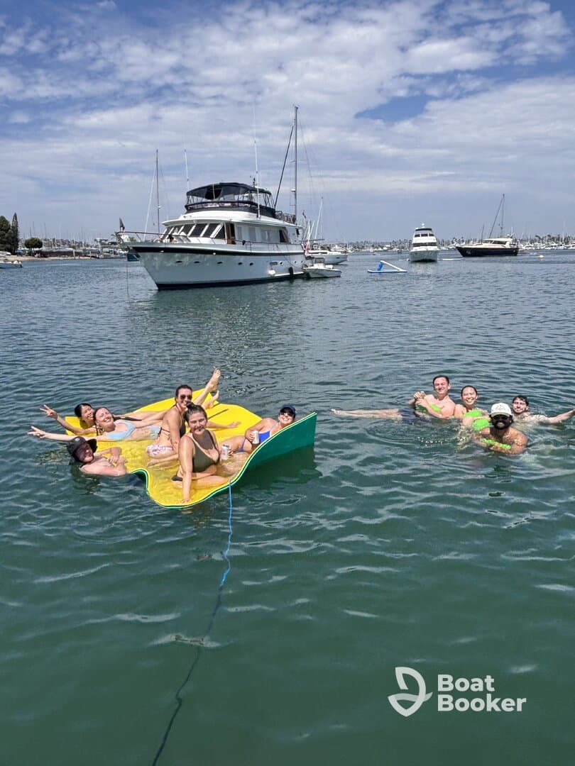 A lively group enjoys a fun day on the water, relaxing on a floating mat while swimming near a yacht — perfect for summer parties and aquatic adventures.