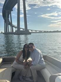 A couple enjoys a sunny day on a boat, smiling together with a scenic bridge in the background — perfect for romantic outings or special celebrations on the water.