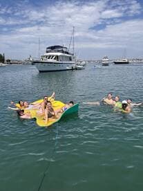 A lively group enjoys a fun day on the water, relaxing on a floating mat while swimming near a yacht — perfect for summer parties and aquatic adventures.