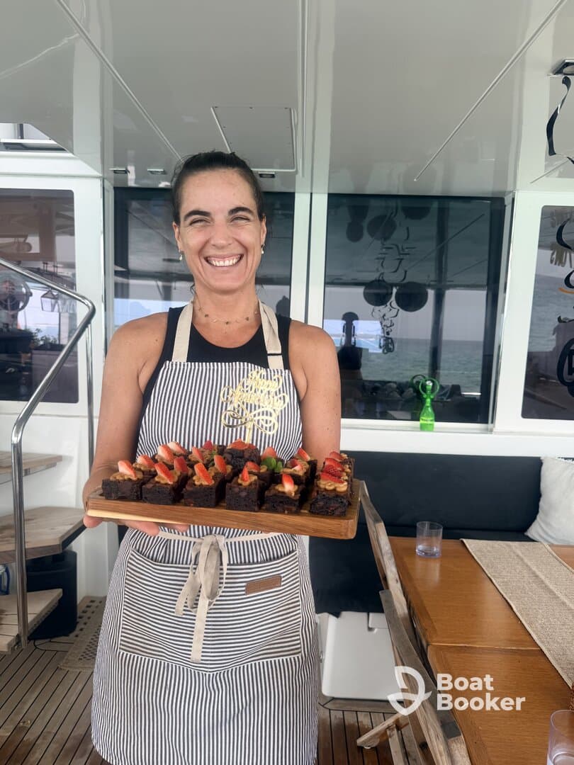 A smiling chef presents a beautifully arranged platter of brownies topped with strawberries aboard a luxurious yacht, showcasing the culinary delights available for guests.