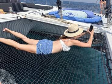 A woman relaxes on the netting of a catamaran, capturing the moment on her phone while enjoying the sun and clear waters — a perfect snapshot of leisure on the water.