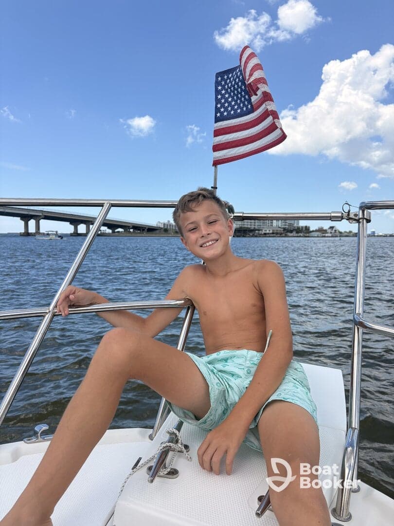 A young boy smiles while sitting on the bow of a boat, with an American flag waving in the background — a perfect moment capturing the joy of a day on the water.