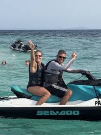 A joyful couple enjoys a thrilling ride on a jet ski in clear waters, surrounded by friends swimming nearby — perfect for adventurous water activities.