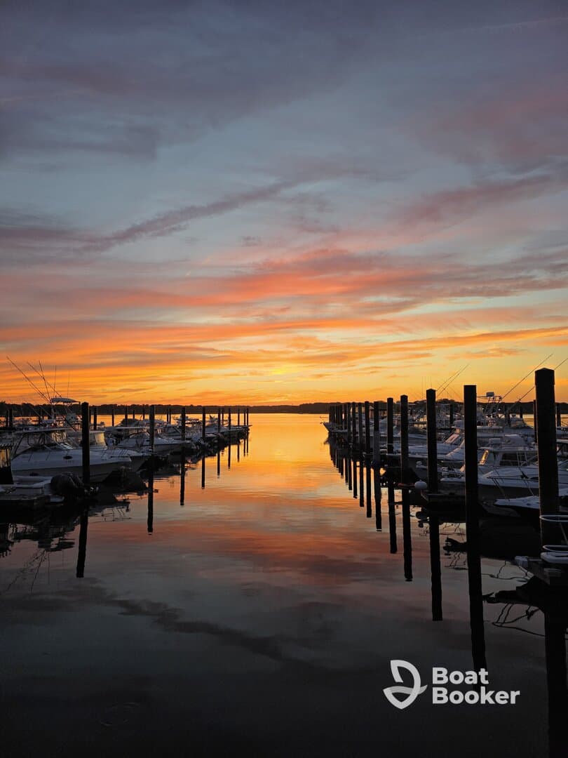 A stunning sunset reflects off the calm waters of a marina, showcasing a row of boats docked peacefully — perfect for evening strolls or romantic getaways.