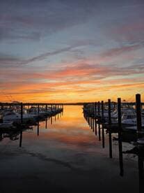 A stunning sunset reflects off the calm waters of a marina, showcasing a row of boats docked peacefully — perfect for evening strolls or romantic getaways.