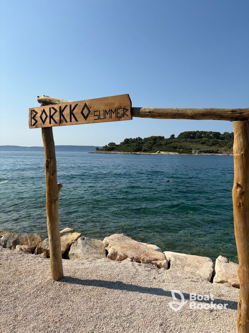 A wooden sign reading 'BORKKO SUMMER' stands by the water's edge, inviting visitors to enjoy the scenic beach and clear waters.